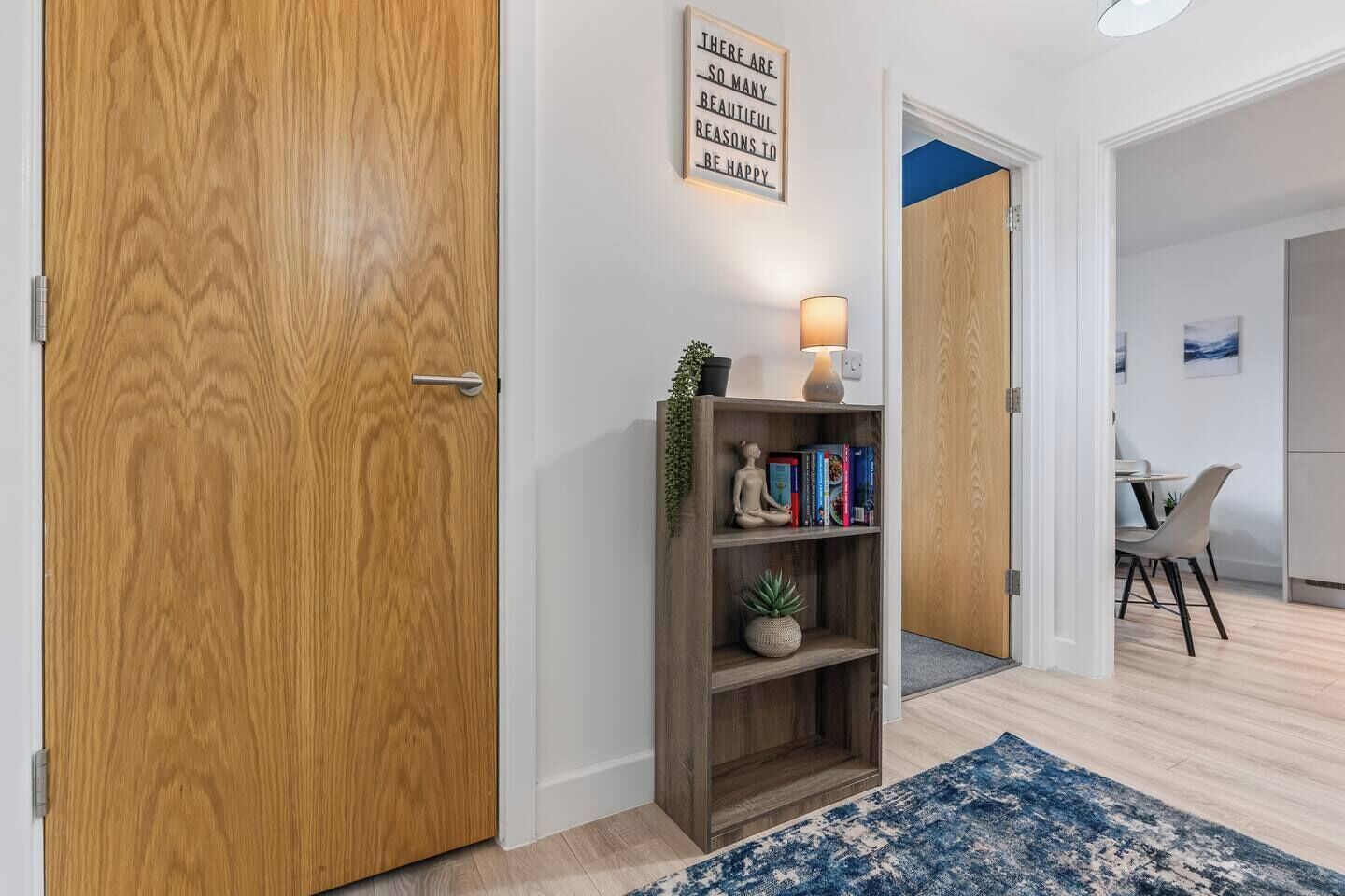 Een gang met een houten deur, een bord aan de muur, een plank met boeken en een plant, en een blauw vloerkleed.