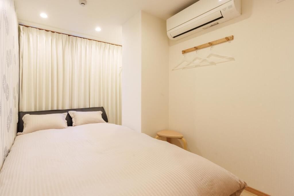 A hotel room with a bed, a small stool, an air conditioning unit, and a wall-mounted rack.