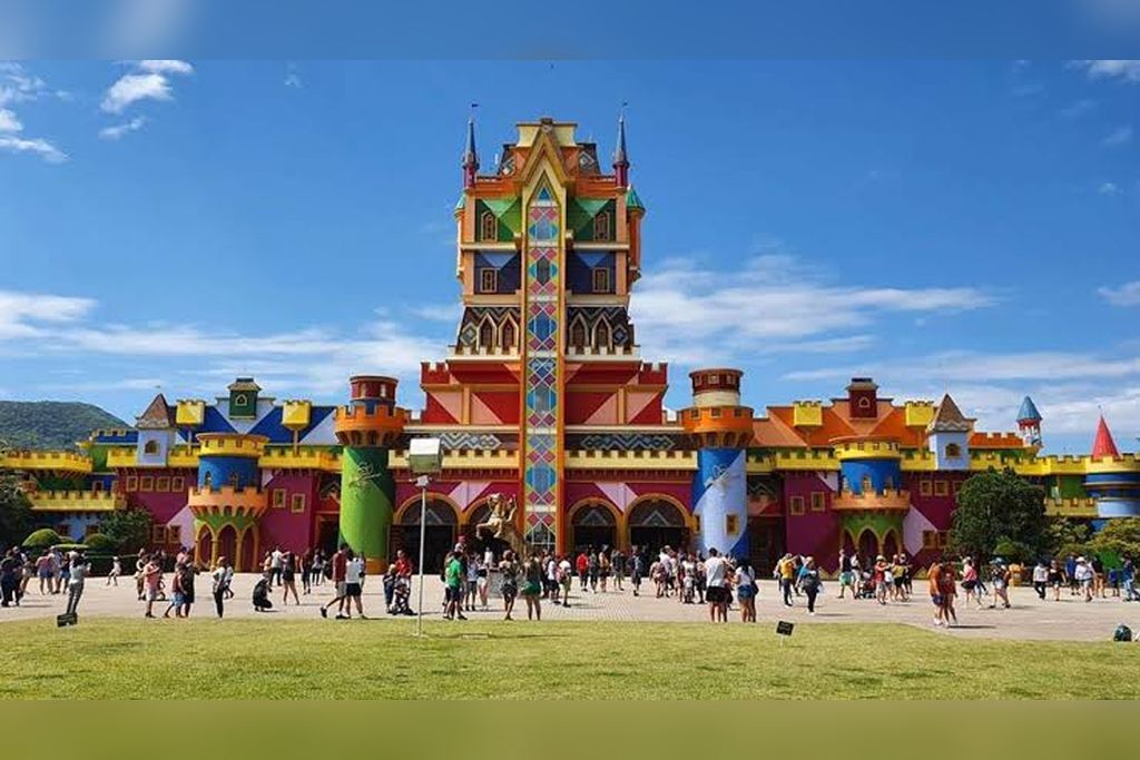 A colorful castle-like structure with multiple towers and a central spire, surrounded by a grassy area with people.