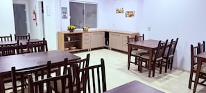 Un espace repas avec plusieurs tables et chaises en bois, un coin cuisine avec des placards et un plan de travail, et un extincteur fixé au mur.