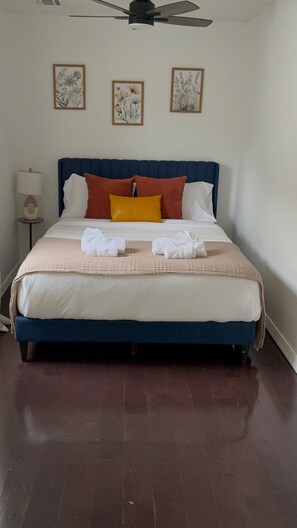 A neatly made bed with a blue headboard, white and orange pillows, and a beige blanket. There are three framed floral artworks on the wall above the bed.