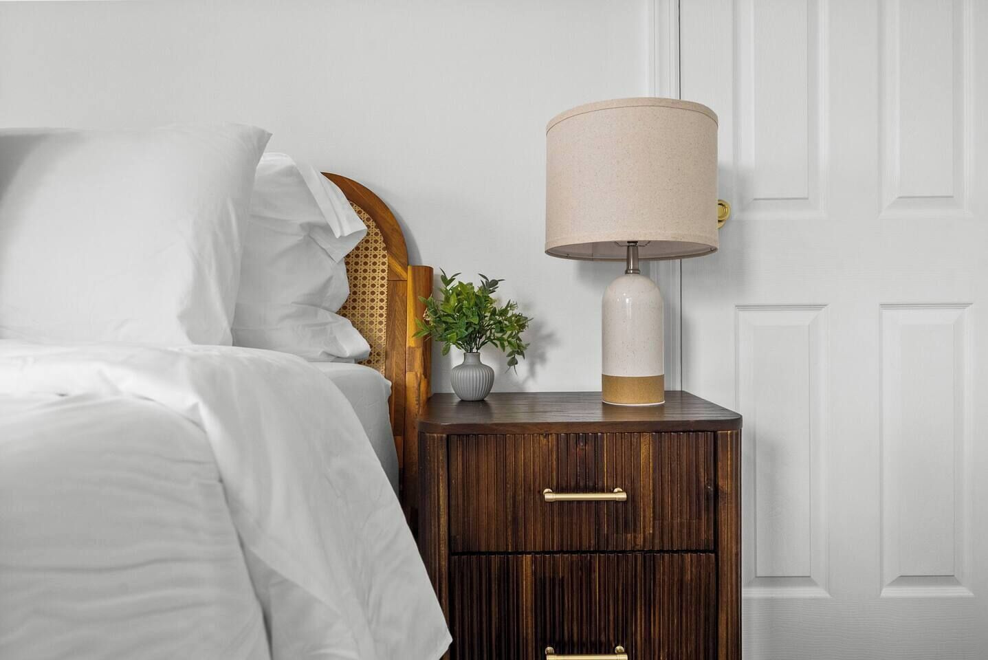Un lit bien fait, avec du linge de lit blanc, une table de chevet en bois sur laquelle se trouve une lampe, et une petite plante en pot.
