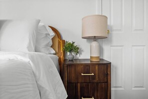A neatly made bed with white linens, a wooden nightstand with a lamp, and a small potted plant.
