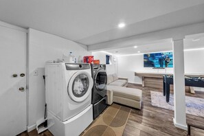 A laundry room with a washing machine, dryer, and ironing station. There is a sofa and a television in the living area.