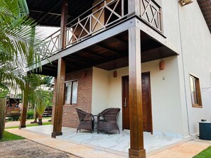 Uma área externa coberta com varanda de madeira, duas cadeiras de vime e uma porta.
