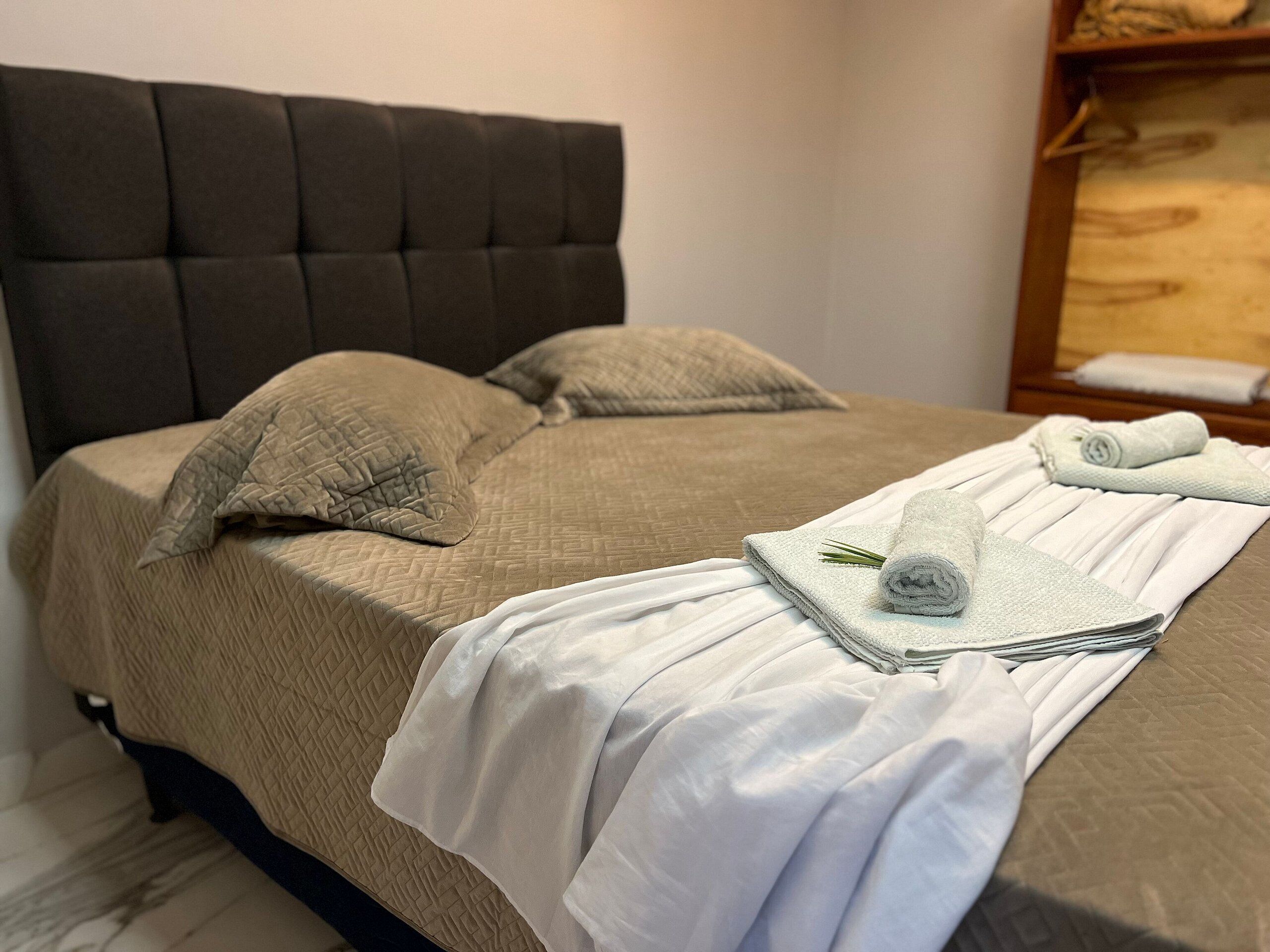 Cama bem arrumada com cabeceira, travesseiros e toalhas dobradas.