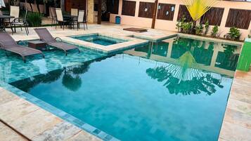 Piscina com espreguiçadeiras e uma mesa, rodeada por um alpendre com plantas e um edifício ao fundo.