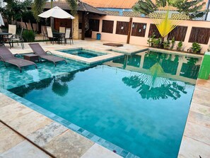A swimming pool with lounge chairs and a table, surrounded by a patio area with plants and a building in the background.