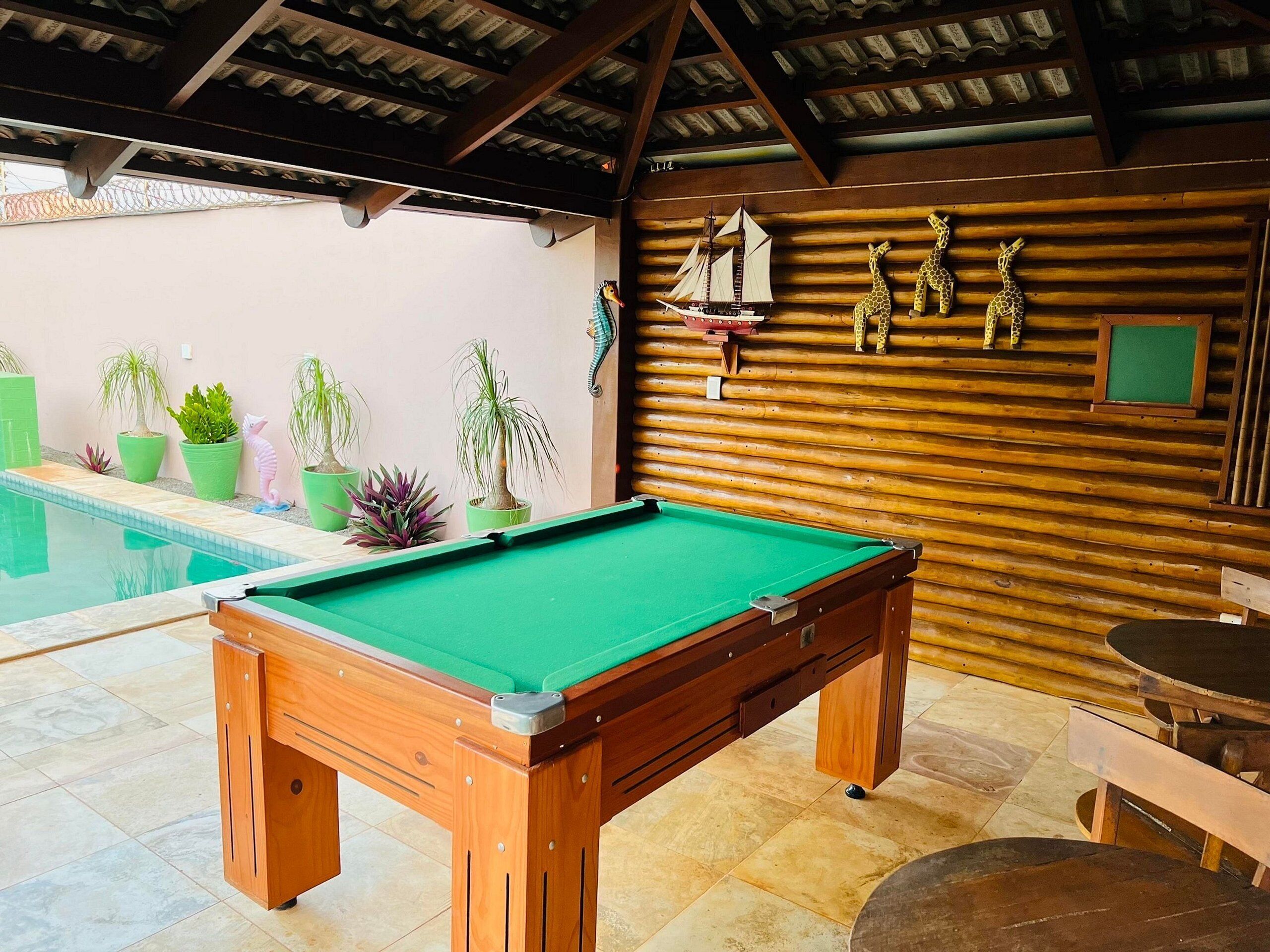 Uma mesa de bilhar com estrutura de madeira e superfície verde aveludada, cercada por plantas em vasos e um pequeno modelo de navio na parede.
