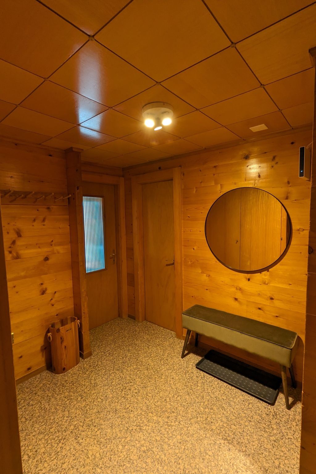 Une pièce avec un plancher en bois, un banc, une porte et un miroir rond accroché au mur.