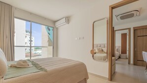 Habitación de hotel con cama, ventana grande, unidad de aire acondicionado, espejo y puerta que da a otra habitación.