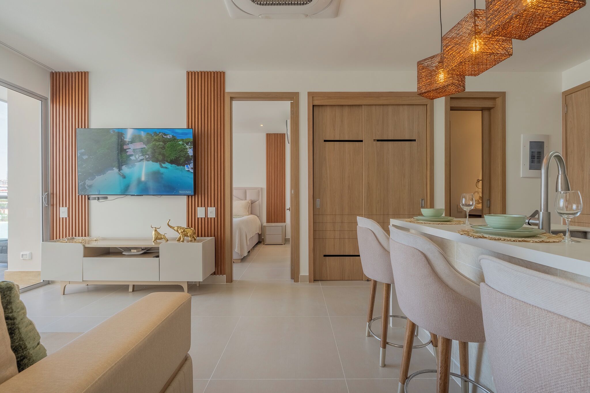 Una sala moderna con televisor de pantalla plana, un sofá, una zona de comedor con taburetes altos y una cocina con pileta de acero inoxidable.