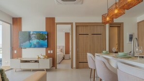Una sala moderna con televisor de pantalla plana, un sofá, una zona de comedor con taburetes altos y una cocina con pileta de acero inoxidable.