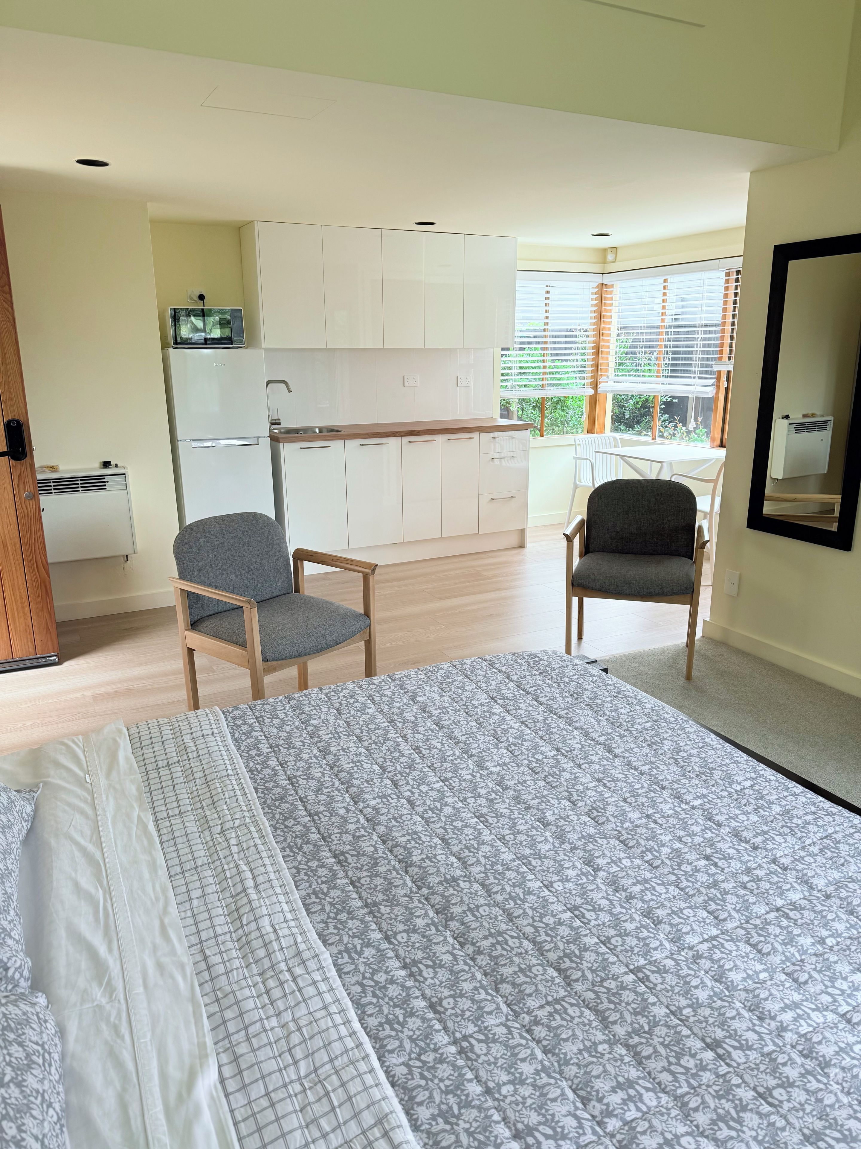 A bedroom with a bed, two chairs, a kitchenette, and a window.