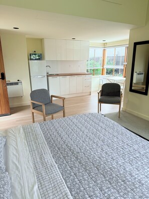 A bedroom with a bed, two chairs, a kitchenette, and a window.