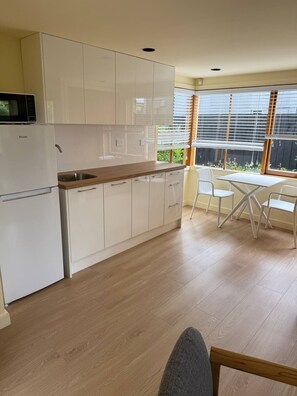 A modern kitchen with white cabinets, a built-in microwave, and a wooden countertop. There is a dining area with a table and chairs, and a window with blinds. - Guesthouse close to beach, Milford Beach, Auckland, New Zealand (Auckland)