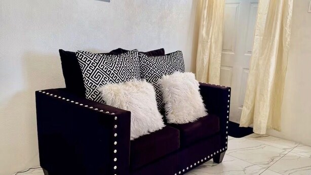 Een donkergrijze bank met decoratieve studs, twee kussens met een patroon, en een lichtgekleurde muur met een stopcontact.