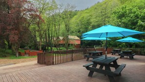 Une terrasse en bois, agrémentée de tables de pique-nique et de parasols bleus, entourée d’arbres et d’une clôture en bois.
