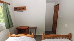 Une petite pièce avec deux lits en bois, une table en bois, une chaise, un radiateur, une fenêtre avec un rideau vert et une porte en bois.