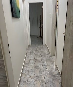 Um corredor estreito com chão de azulejos, paredes brancas e uma porta no final.