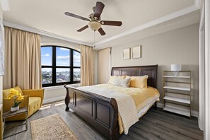 A bedroom with a large bed, a ceiling fan, a window with curtains, a bedside table, and a view of the city. - 3BR Harborfront Condo - Pool, Hot Tub, Bay Shuttle (Destin)