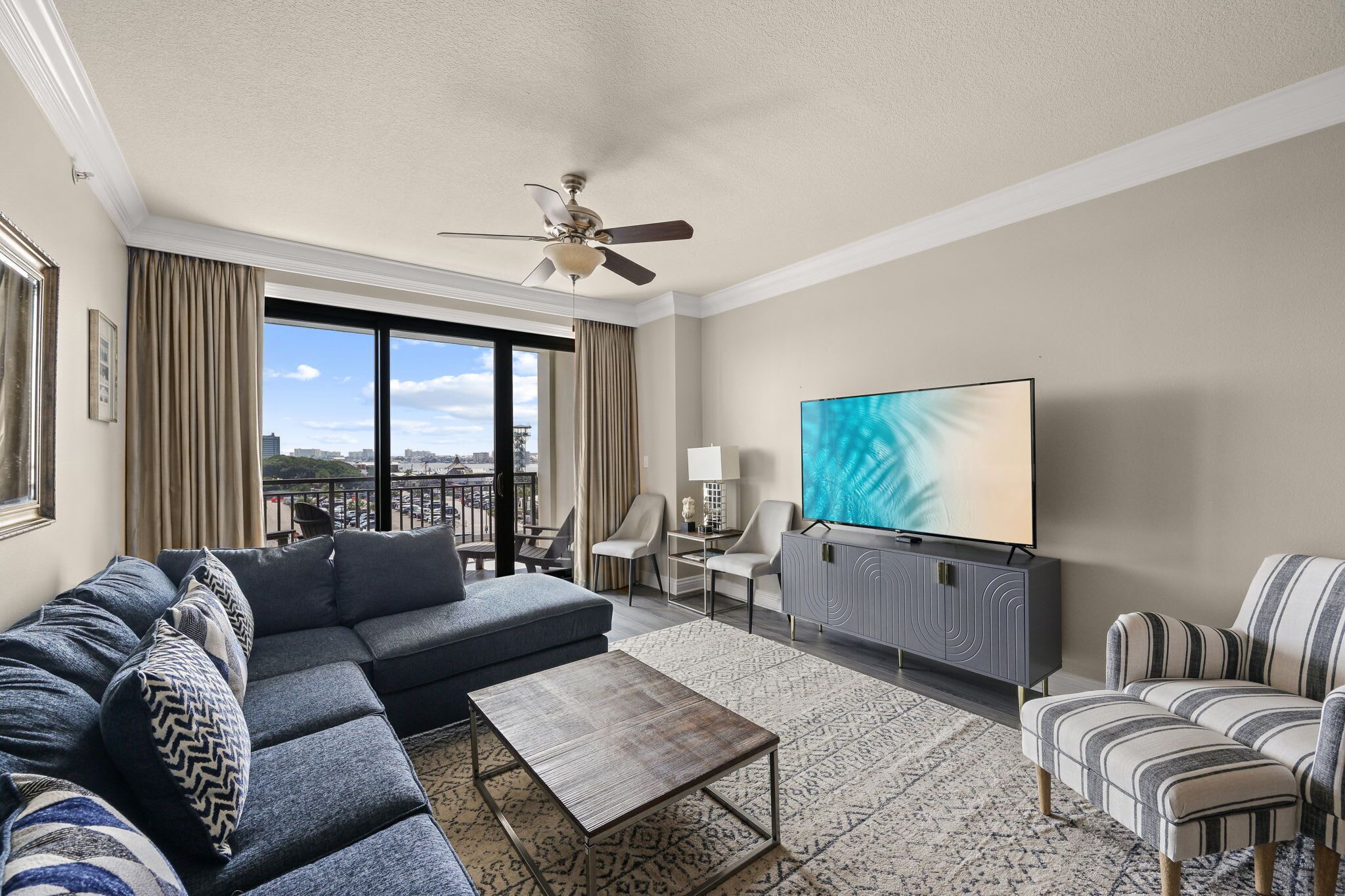 A modern living room with a sectional sofa, a flat-screen TV, a ceiling fan, and a view of the cityscape through large windows.