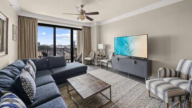 A modern living room with a sectional sofa, a flat-screen TV, a ceiling fan, and a view of the cityscape through large windows. - 3BR Harborfront Condo - Pool, Hot Tub, Bay Shuttle (Destin)