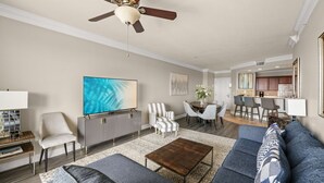 A spacious living room with a ceiling fan, a flat-screen TV, a grey sofa, a coffee table, and a dining area with chairs and a table. - 3BR Harborfront Condo - Pool, Hot Tub, Bay Shuttle (Destin)