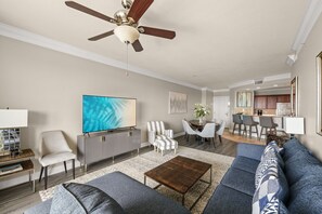 Ein geräumiges Wohnzimmer mit Deckenventilator, einem Flachbildfernseher, einem grauen Sofa, einem Couchtisch und einem Essbereich mit Stühlen und einem Tisch.