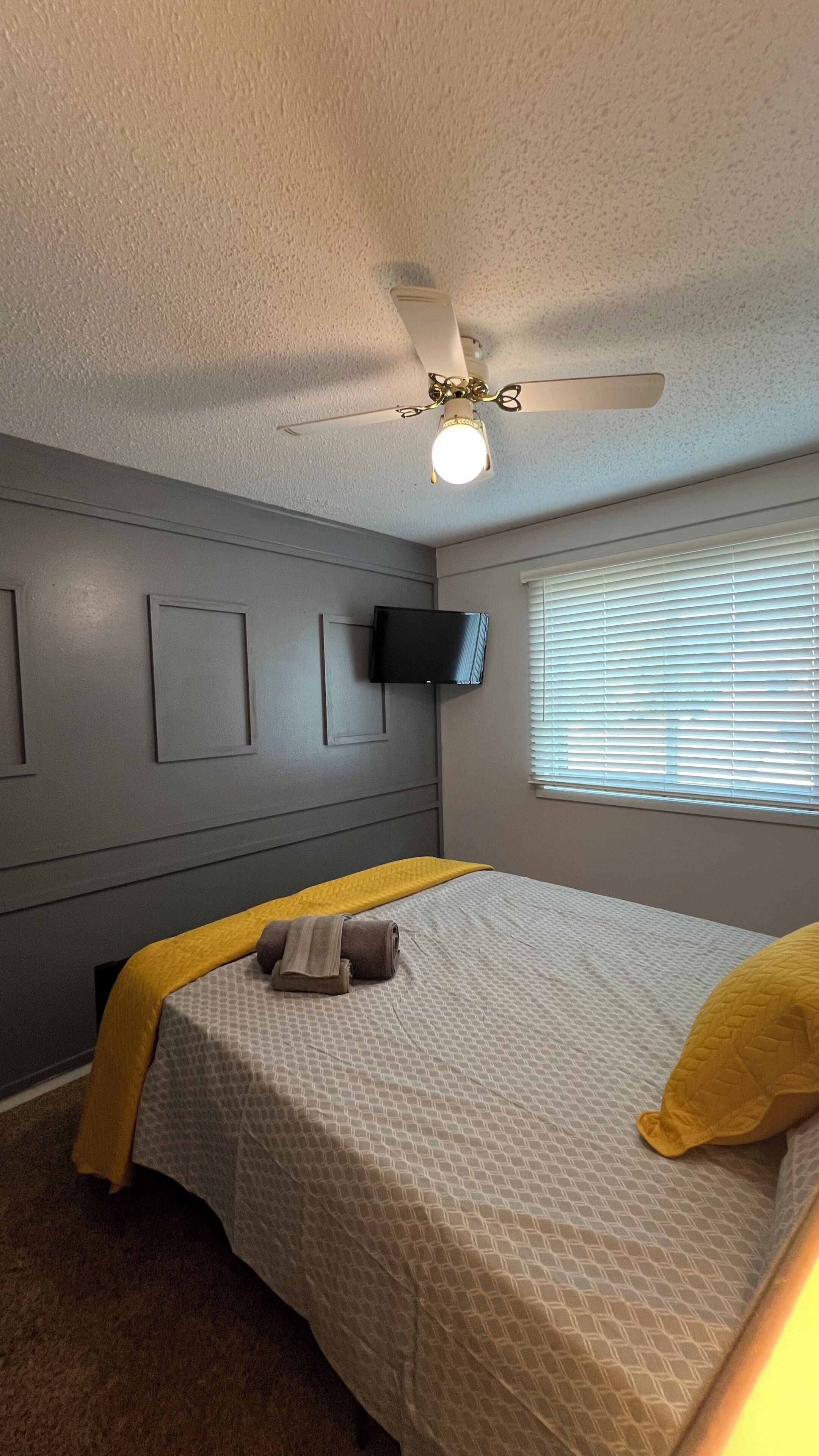 Une chambre à coucher avec un lit, un ventilateur de plafond, un téléviseur fixé au mur et une fenêtre avec des stores.