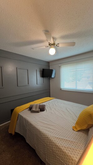 Quarto com cama, ventoinha no teto, TV fixada na parede e janela com persianas.