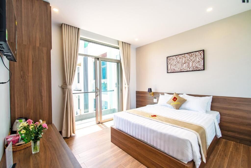 A hotel room with a bed, a wooden headboard, a window with curtains, a wall-mounted artwork, and a small table with flowers.