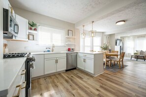 A modern kitchen with white cabinetry, a gas stove, and a dining area with a wooden table and chairs. - 10 Min to Lucas Oil~Walk to Mass Ave~Hot Tub (Indianapolis)