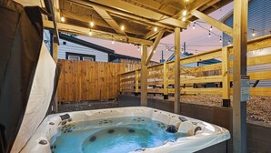 Un jacuzzi bajo una pérgola de madera con luces colgantes, rodeado de cercas de madera.