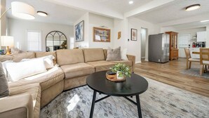 Una sala de estar moderna con un sofá beige, una mesa de centro con una planta, y una cocina al fondo.