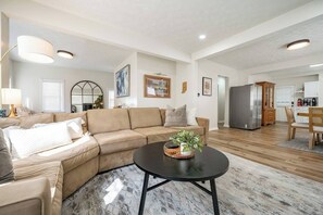 A modern living room with a beige sofa, a coffee table with a plant, and a kitchen in the background. - 10 Min to Lucas Oil~Walk to Mass Ave~Hot Tub (Indianapolis)