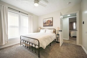 Un dormitorio con cama, mesita de noche, lámpara, un cuadro en la pared y una puerta que da a otra habitación.