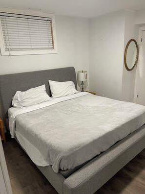 A bedroom with a single bed, a bedside table with a lamp, a window with blinds, and a mirror on the wall.