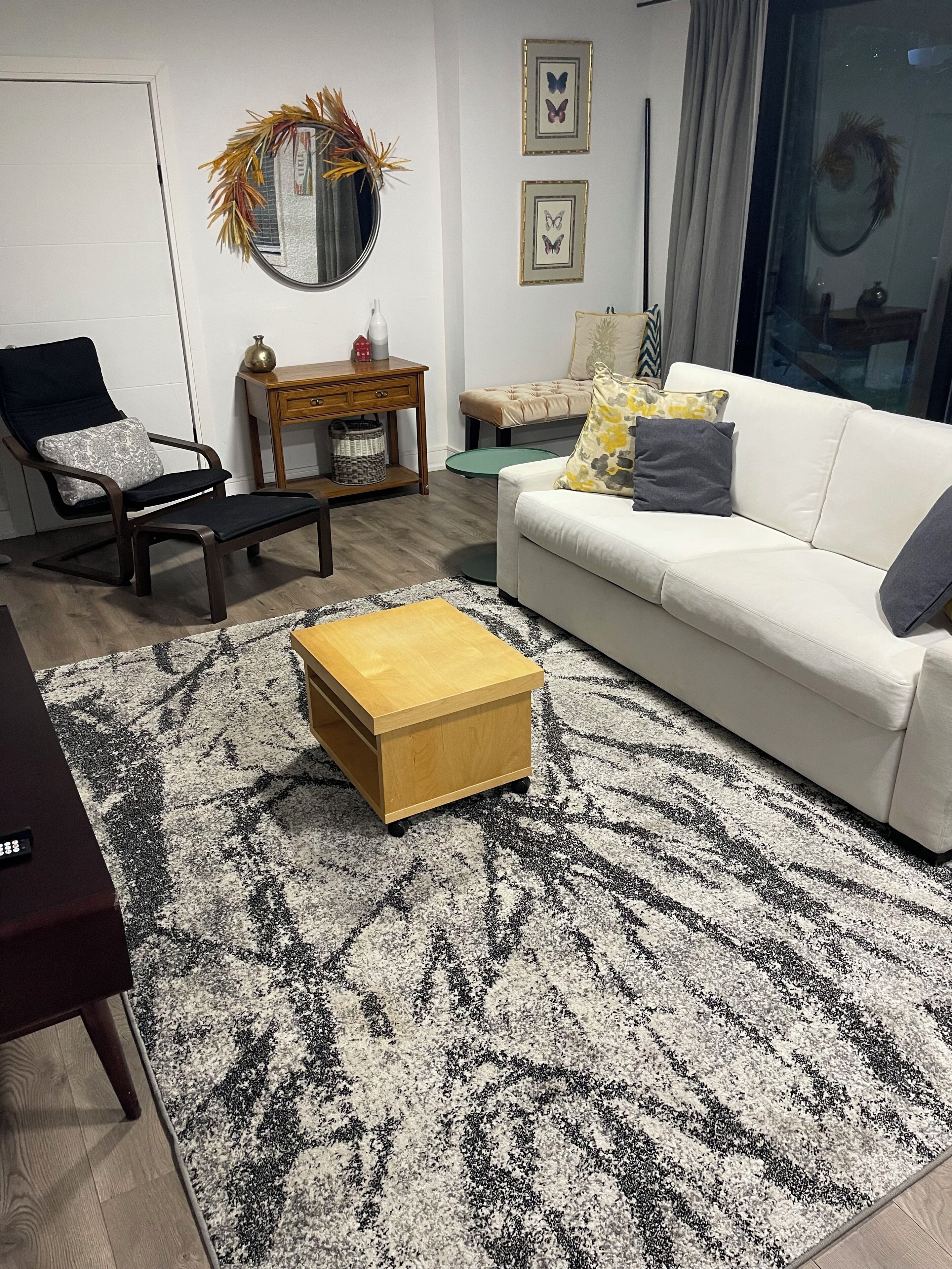 Een woonkamer met een witte bank, een houten salontafel, een zwart-wit tapijt met een patroon, een ronde spiegel en een klein bijzettafeltje.