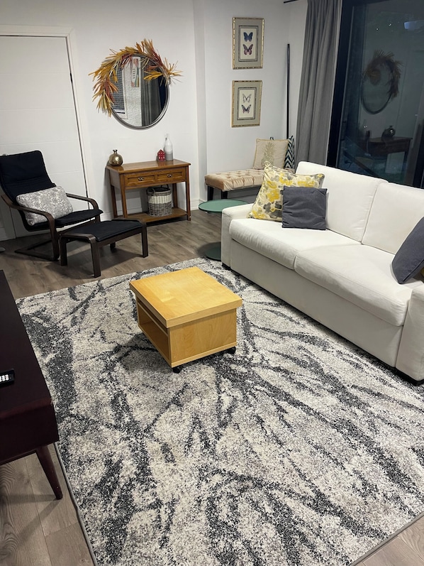 A living room with a white sofa, a wooden coffee table, a black and white patterned rug, a round mirror, and a small side table.