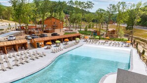 A swimming pool with lounge chairs and umbrellas, surrounded by wooden buildings and a hilly landscape. - The Lazy Beaver Lodge - Hot Tub (Hollister)