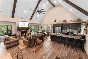 A spacious living area with a fireplace, a dining table, and a kitchen with dark cabinets and stainless steel appliances. - The Lazy Beaver Lodge - Hot Tub (Hollister)