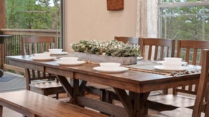 A wooden dining table set with white plates and bowls, flanked by chairs, with a woven basket of greenery on the table. - The Lazy Beaver Lodge - Hot Tub (Hollister)