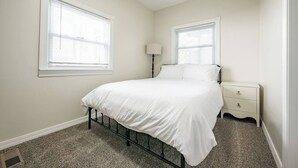 Um quarto com uma cama, duas janelas, uma mesa de cabeceira e uma luminária.