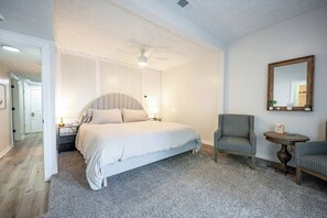 Ein Schlafzimmer mit einem Bett, einem Sessel, einem kleinen Tisch, einem Spiegel und einer Tür zu einem anderen Raum.