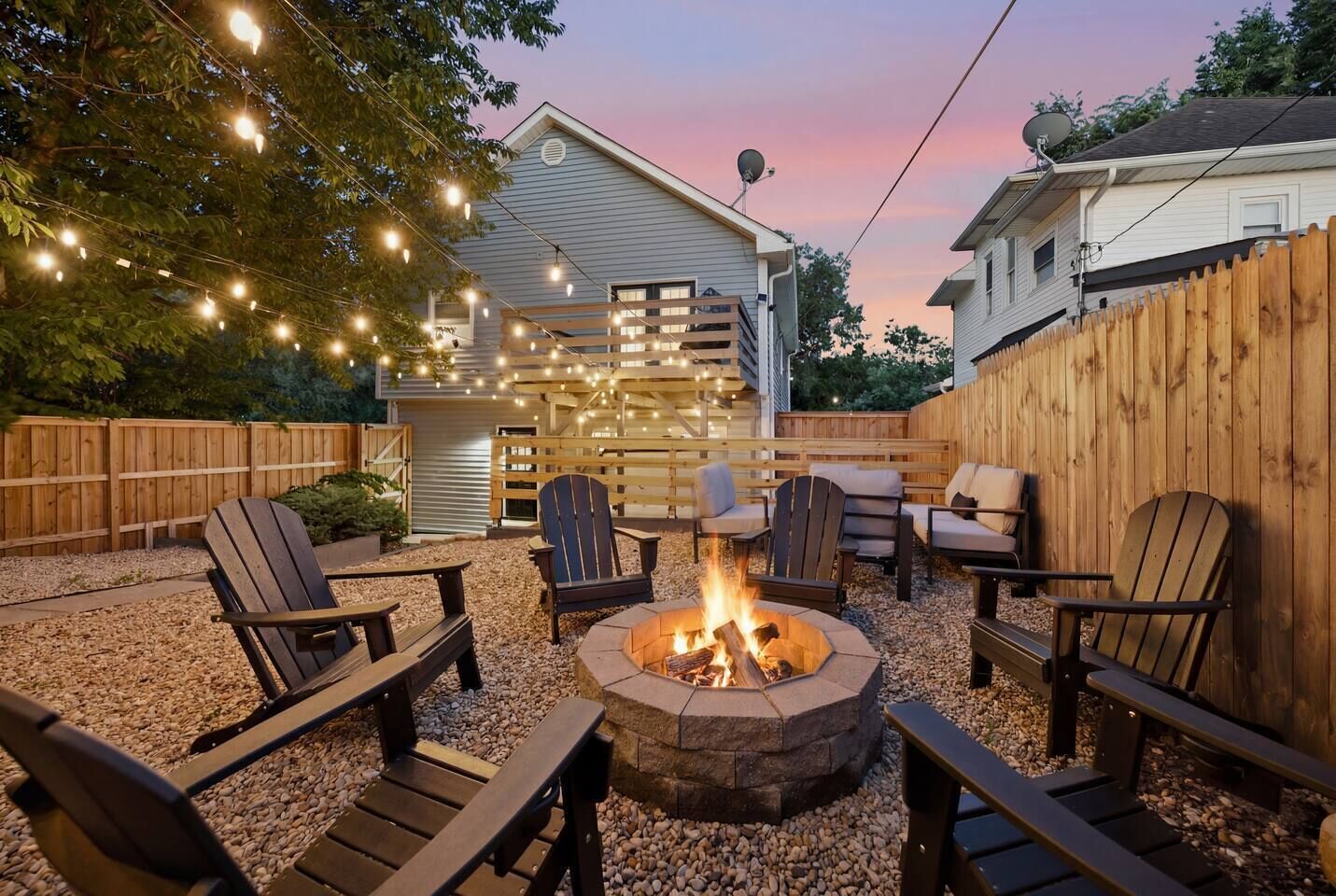 A backyard with a fire pit, outdoor seating, string lights, and a wooden fence.