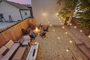 Ein Garten mit Feuerstelle, umgeben von Gartenmöbeln, einem Holzzaun und Lichterketten.