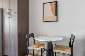 Pieni ruokailutila, jossa on pyöreä pöytä ja kaksi tuolia, kaappi sekä seinällä oleva kehystetty abstrakti maalaus.