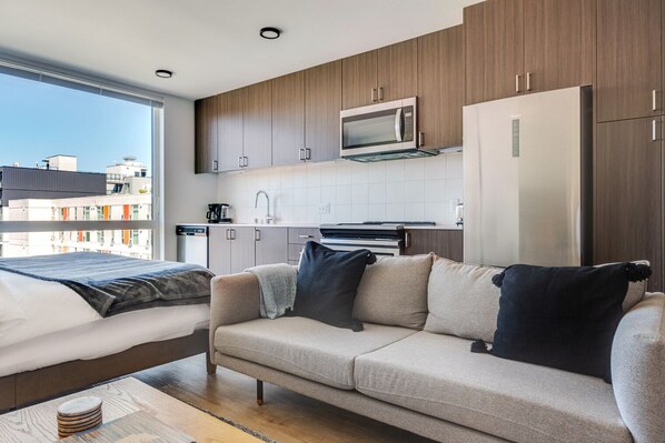 Moderni hotellihuone, jossa on pieni keittiönurkkaus, tyynyillä varustettu sohva, peitolla varustettu sänky ja näkymä kaupunkimaisemaan.
