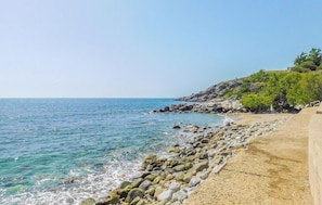 Una playa costera con agua azul cristalina, costa rocosa y un sendero arenoso que lleva al mar.